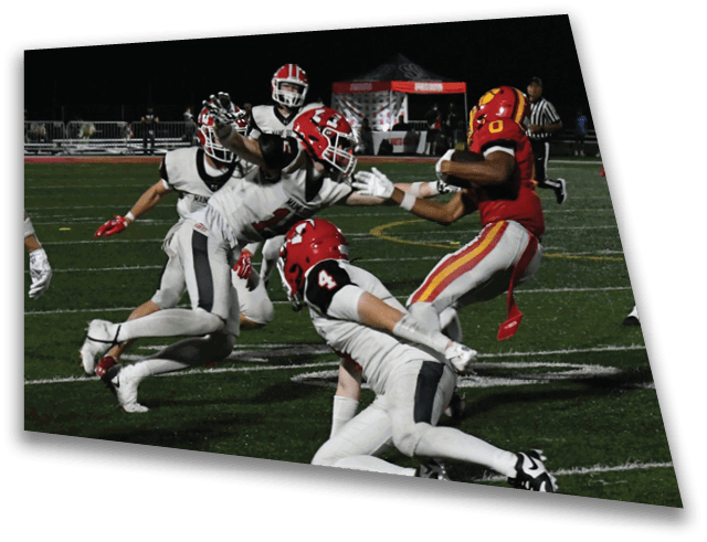A group of people in football uniforms are actively playing on a field at night with one person in red running with the ball while others in white attempt to tackle. Description generated by AI