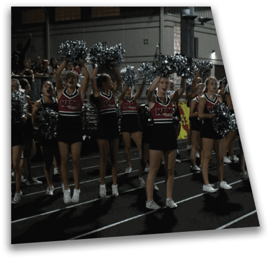 A group of adults in matching cheerleading uniforms with pom-poms are performing on a track at night. Description generated by AI