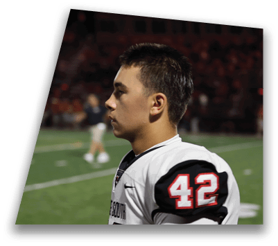A person in a sports uniform stands on the sidelines of a football field with a blurred background of another person and spectators. Description generated by AI
