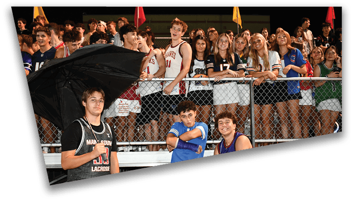 A group of people in sports jerseys and casual clothing are gathered behind a fence with three people in the foreground, one holding a large black umbrella. Description generated by AI