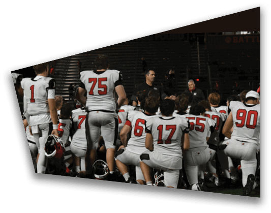 A group of people wearing white sports uniforms with red numbers are kneeling on a field while listening to two standing adults speaking to them. Description generated by AI