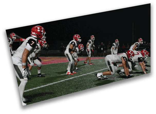 A group of people wearing football uniforms and helmets are positioned on a field at night, preparing for a play. Description generated by AI