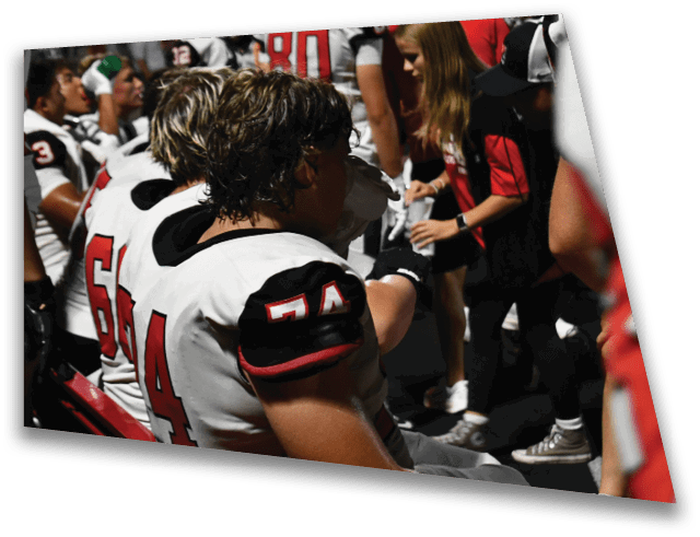 People wearing football uniforms are sitting on a bench while others stand nearby, possibly during a game or practice. Description generated by AI