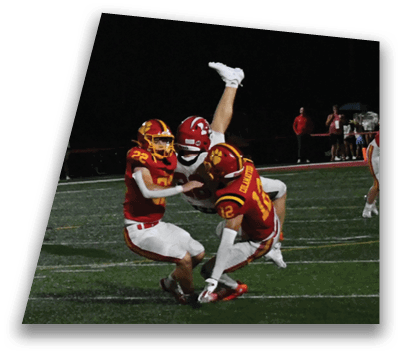 Three football players in red and yellow uniforms are tackling a player in a white uniform who is upside down on a football field at night. Description generated by AI
