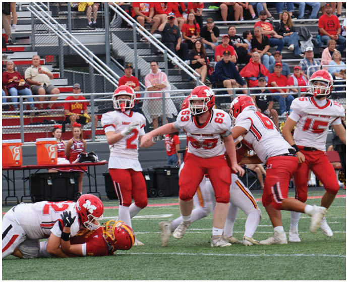 A group of football players in red and white uniforms are on a field with one player tackling another near the ground while spectators watch from bleachers in the background. Description generated by AI