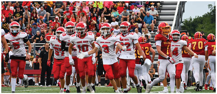 A group of people in red and white football uniforms are running on a green field with spectators in the background. Description generated by AI