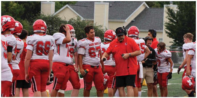 A group of people wearing red sports uniforms and cleats stand on a grassy field near sports equipment and bags. Description generated by AI