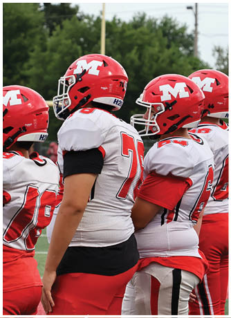 Two people wearing red and white football uniforms and helmets stand on a football field near a person holding an orange down marker. Description generated by AI