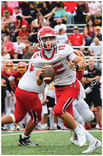 A person in a red and white football uniform is holding a football and preparing to throw it during a game with other players and spectators in the background. Description generated by AI