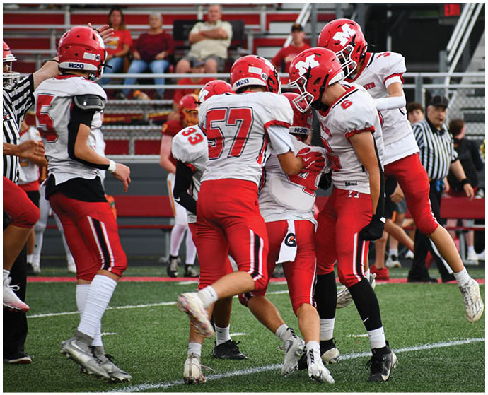 Four people in red and white football uniforms are celebrating on a football field with a referee and cheerleaders in the background. Description generated by AI
