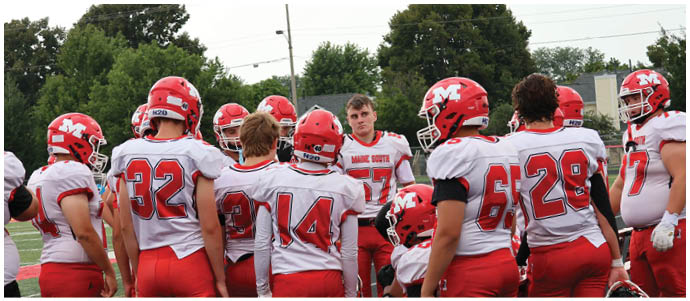A group of people wearing red and white football uniforms and helmets are gathered closely together outdoors, appearing to be in a team huddle or discussion. Description generated by AI