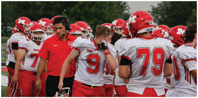 A group of people wearing red and white football uniforms and helmets are gathered on a field with one person in a red shirt and headset appearing to coach or direct them. Description generated by AI