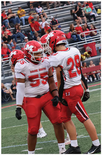 Two people in red and white football uniforms stand on a green field near a sideline with seated cheerleaders and spectators in the background. Description generated by AI