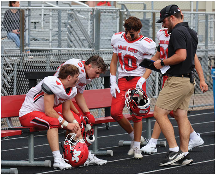 Three people in red and white football uniforms are sitting and standing near a bench on a track while another person in a black shirt and khaki shorts holds a tablet nearby. Description generated by AI