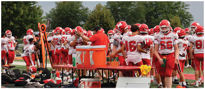 Three people in red and white football uniforms stand on a green field with others practicing in the background. Description generated by AI