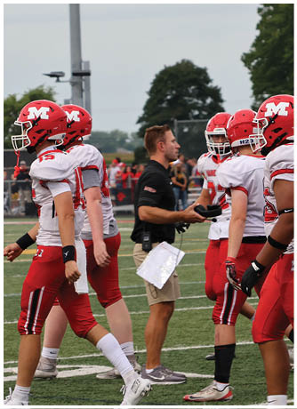 A group of people wearing red and white football uniforms and helmets stand on a field while a person in a black shirt and khaki shorts appears to be giving instructions. Description generated by AI