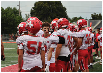 A group of people wearing red and white football uniforms and helmets are gathered on a field, with one person facing forward showing black face paint. Description generated by AI