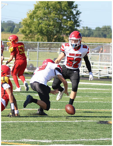 Two people in sports uniforms are on a football field with one person kicking a football while others stand in the background near bleachers. Description generated by AI