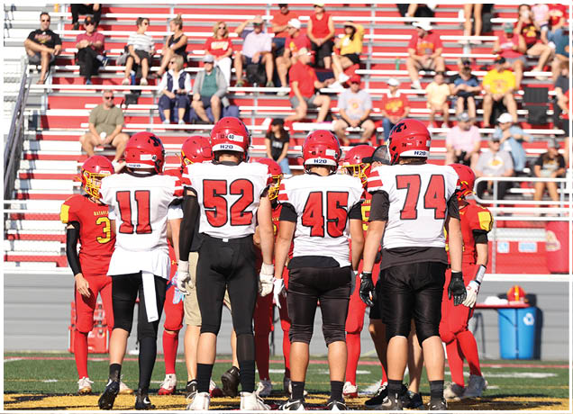 A group of people wearing football uniforms are standing together on a field with spectators seated in bleachers behind them. Description generated by AI