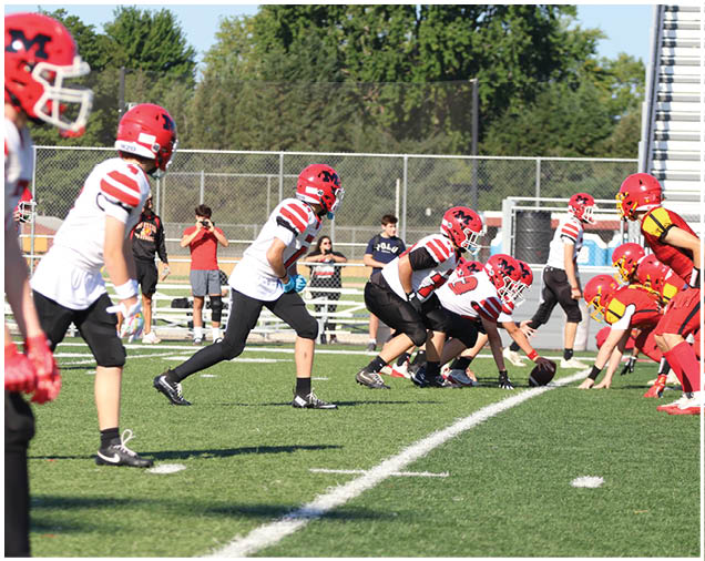 Players in red and white and red and yellow football uniforms are lined up on a green field preparing for a play during a game. Description generated by AI