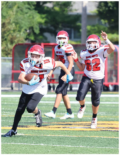 Four people wearing football uniforms are on a field with one person running while holding a football and others running behind. Description generated by AI