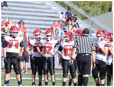 A group of people in football uniforms and helmets stand on a field with a referee in a striped shirt facing them. Description generated by AI