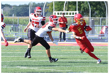 Adults wearing red and white football uniforms are playing on a green field with one player in red running with the ball while others in white attempt to block or tackle. Description generated by AI