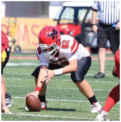 A group of people in football uniforms are on a field with one person in a white and red uniform preparing to snap the ball while others in red and yellow uniforms are positioned to defend. Description generated by AI