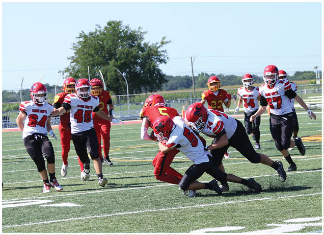 Players in red and white football uniforms are actively engaged in a tackle on a green field with a clear sky and trees in the background. Description generated by AI