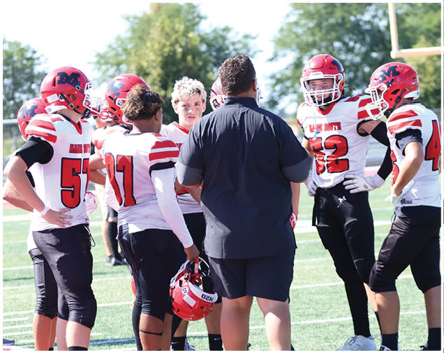 A group of people wearing football uniforms and helmets are gathered on a field with one person in a black shirt and shorts appearing to give instructions. Description generated by AI