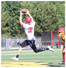 A person wearing a red helmet and white jersey with the number 2 is jumping to catch a football on a grassy field while a referee in a striped shirt watches nearby. Description generated by AI