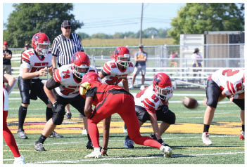 Several people in football uniforms are positioned on a field with one person in red preparing to snap the ball to a player in white and red. Description generated by AI