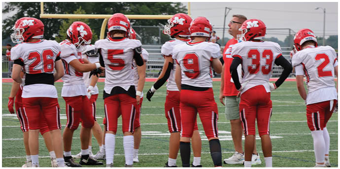 A group of people wearing red and white sports uniforms stand on a green field with their backs facing the camera. Description generated by AI