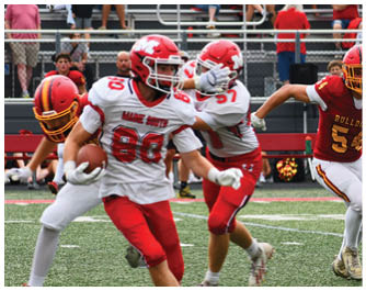 People wearing football uniforms are running on a grassy field during a game, with one player in a red and yellow “Bulldogs" jersey and others in white and red jerseys. Description generated by AI