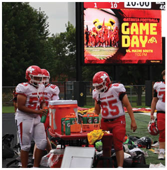 People in red and white football uniforms are gathered on the sidelines of a football field with equipment and water coolers nearby. Description generated by AI