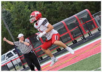 A person in red and white football gear is jumping near a referee who is signaling with a thumbs-up on a football field. Description generated by AI