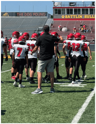 A group of people wearing red helmets and white jerseys with black pants are gathered on a football field near a coach in black shirt and shorts, with empty bleachers and a scoreboard in the background. Description generated by AI