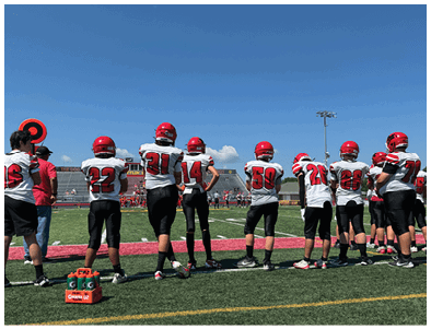 A group of people wearing red helmets and white sports jerseys stand on the sidelines of a football field under a clear blue sky. Description generated by AI