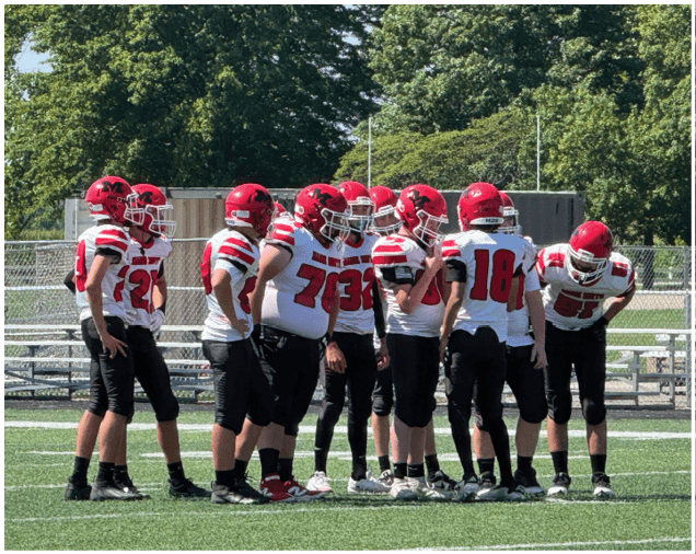 A group of people wearing red helmets and white sports uniforms are gathered outdoors on a sunny day with trees in the background. Description generated by AI