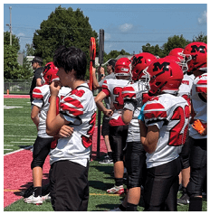 A group of people wearing red helmets and white sports jerseys are standing together on a green field with red markings. Description generated by AI