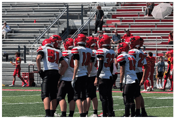 A group of people wearing red helmets and white sports jerseys are gathered on a football field with empty bleachers and a few seated spectators in the background. Description generated by AI