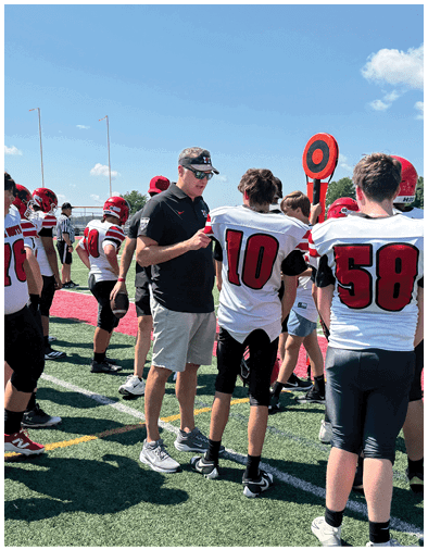 A group of people wearing football uniforms and helmets stand on a field with one person in a black shirt and shorts talking to a player wearing number 10. Description generated by AI