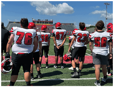 Adults wearing football uniforms stand on a field facing away from the camera toward empty bleachers under a partly cloudy sky. Description generated by AI