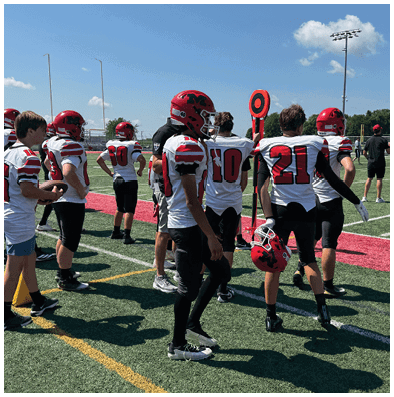 A group of people wearing football uniforms and helmets stand on a turf field near a yard marker during a game or practice. Description generated by AI