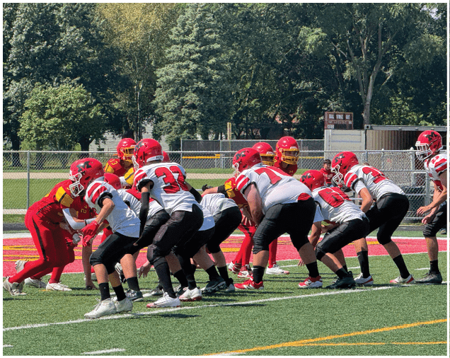 Adults in red and white football uniforms are lined up on a green field preparing for a play during a game. Description generated by AI