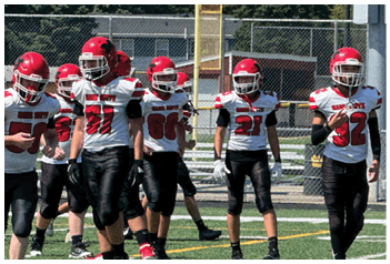 A group of people wearing red helmets and white jerseys with black pants are standing on a football field near a chain-link fence. Description generated by AI