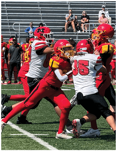 A group of people in red and white football uniforms are playing on a green field with a crowd of people in red uniforms watching from the sidelines and empty bleachers in the background. Description generated by AI