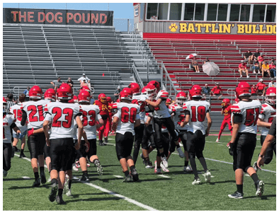 Football players in red and white uniforms are gathered on a field with mostly empty bleachers and a few people sitting under umbrellas in the background. Description generated by AI