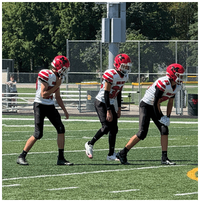 Four people wearing red helmets and black pants are positioned on a football field with a chain-link fence and trees in the background. Description generated by AI