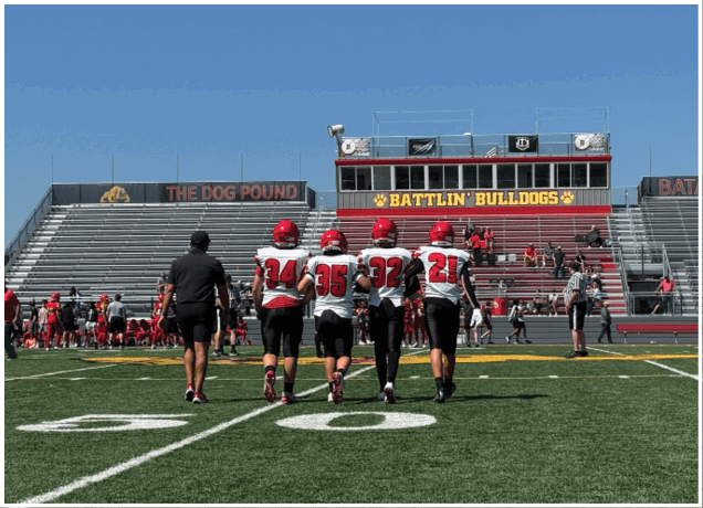 Five people in football uniforms walk together on a green field toward empty bleachers under a clear blue sky. Description generated by AI